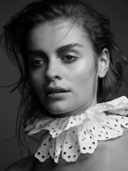 Black and white portrait of a woman with a neutral expression wearing a white, frilled, eyelet lace collar against a dark background.