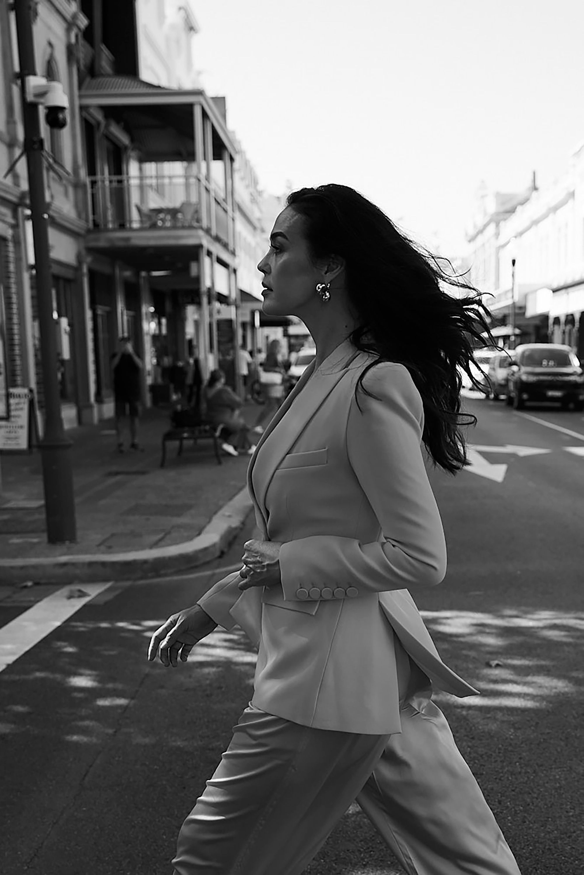 A woman with long dark hair walking down a city street wearing a tailored suit jacket and pants.