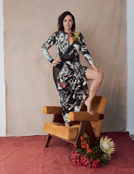 Brunette woman in long-sleeved printed dress poses with one leg on a mustard yellow armchair, with flowers on a red backdrop.