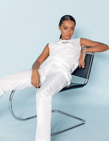 A young adult female in a white sleeveless shirt and pants, seated on a black and chrome chair against a blue background in a fashion shot.