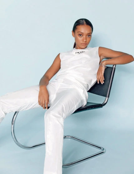 A young adult female in a white sleeveless shirt and pants, seated on a black and chrome chair against a blue background in a fashion shot.