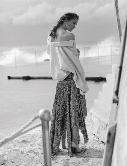 A woman in a shimmering gown and draped top stands near the ocean. The black and white image captures a serene, fashionable moment.