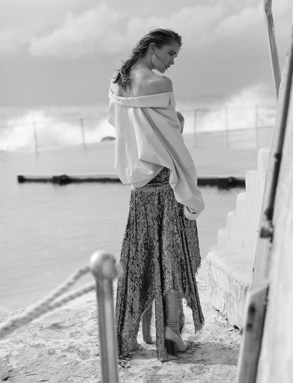 A woman in a shimmering gown and draped top stands near the ocean. The black and white image captures a serene, fashionable moment.