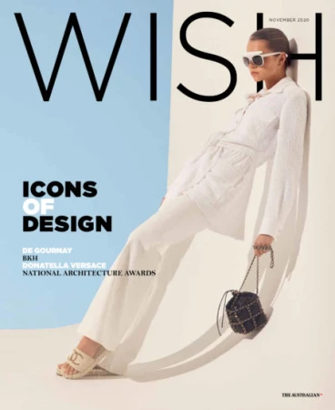 Model in white belted outfit leaning against a curved studio wall holding a black studded handbag