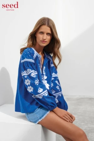 Model seated on a white block wearing a blue embroidered blouse and denim shorts against a white background