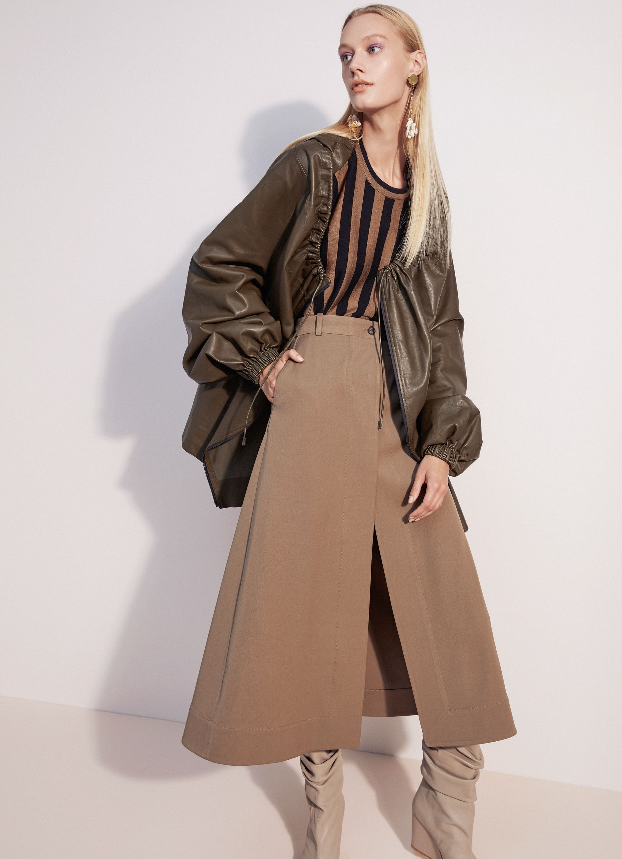 A woman in a green jacket, striped top, and brown skirt poses against a plain white backdrop in a fashion studio shot.