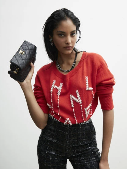 Brunette woman poses indoors wearing a red Chanel sweater, black pants, jewelry, holding a quilted leather purse with gold hardware.