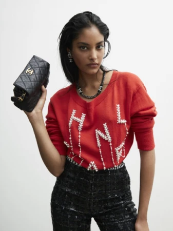 Brunette woman poses indoors wearing a red Chanel sweater, black pants, jewelry, holding a quilted leather purse with gold hardware.