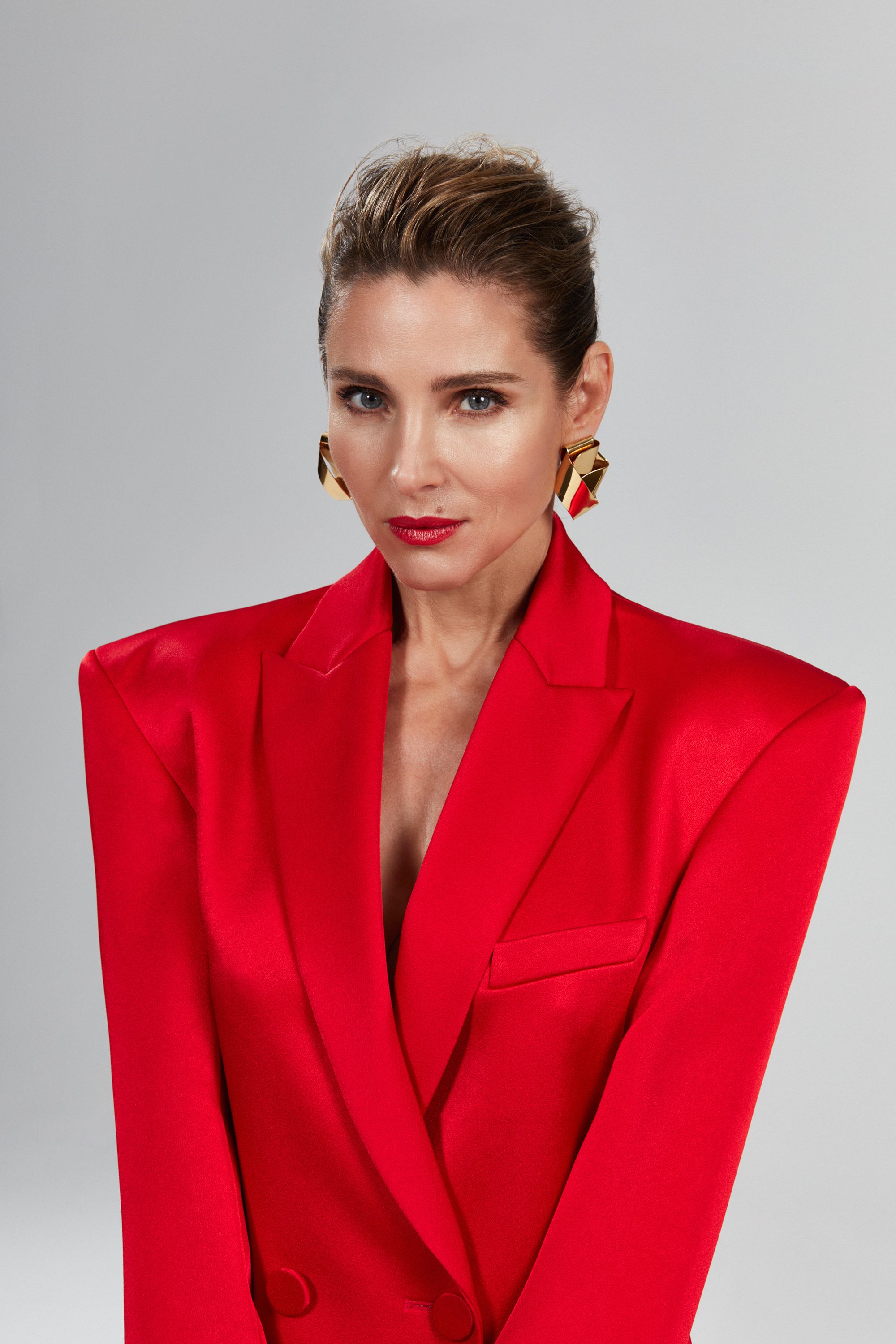 Portrait of an adult woman in a bright red blazer and large gold earrings against a grey studio backdrop. She has a neutral expression.