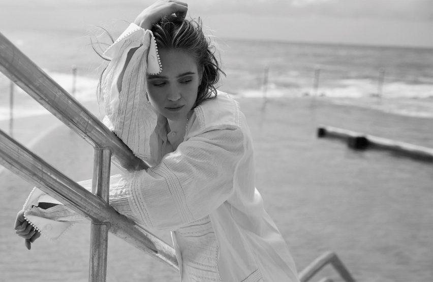 Black and white portrait of a young woman in a textured white tunic leaning on silver railings with the ocean in the background.
