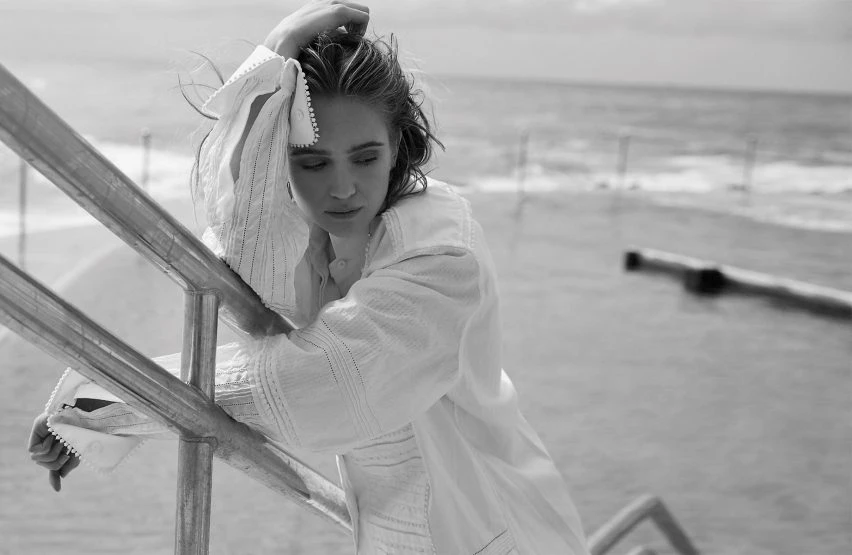 Black and white portrait of a young woman in a textured white tunic leaning on silver railings with the ocean in the background.