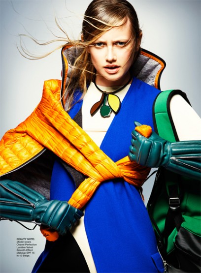 Fashion photo shoot features a woman in a blue dress with orange puffy sleeves and green gloves, set against a light grey background.