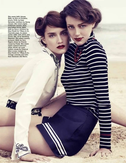 Two adult women pose on a sandy beach wearing fashionable summer clothing with dark red lipstick and styled hair. Ocean visible.