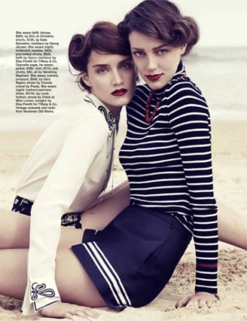 Two adult women pose on a sandy beach wearing fashionable summer clothing with dark red lipstick and styled hair. Ocean visible.