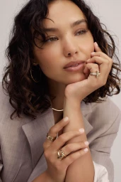 Close-up portrait of a woman with curly dark hair wearing a grey blazer, gold rings, and hoop earrings, styled by Brad Mullins.