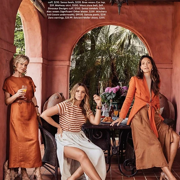 Three women model rust, brown, and cream-colored clothing with a garden and arched doorway in the background, outdoor fashion photography.