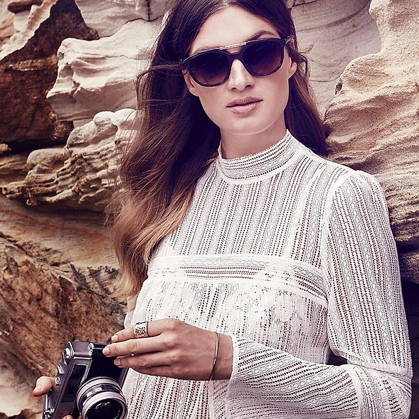 Adult female posing outdoors wearing sunglasses and a long-sleeve white dress while holding a vintage camera near rock formations.