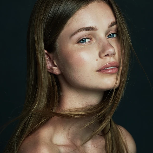 Close-up portrait of a young, light-skinned woman with long brown hair posing against a dark gray backdrop, showcasing beauty and fashion.