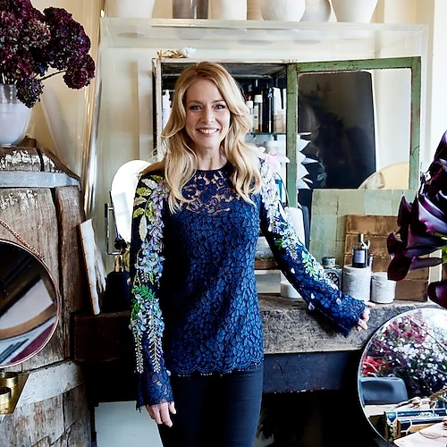 Blonde woman in navy blue lace top with floral embellishments stands indoors surrounded by vases, mirrors, and rustic decor.