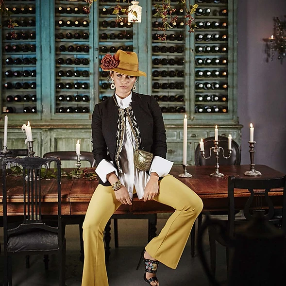 Stylish woman in hat and mustard-yellow bell-bottoms poses indoors near dining table and wine rack, capturing a blend of elegance and retro charm.