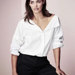Woman in an off-the-shoulder white shirt and dark trousers posing against a pale background
