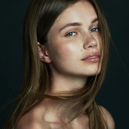 Close-up portrait of a young woman with long brown hair, blue eyes, and natural makeup against a dark background.