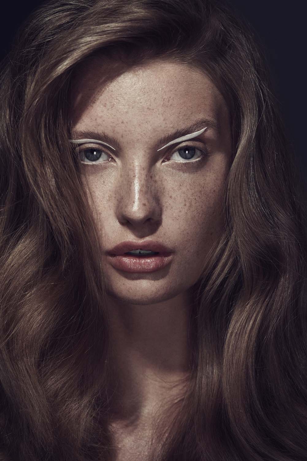 Close-up beauty shot of a freckled woman with long brown wavy hair, bold white eyeliner, and natural lip color against a dark background.