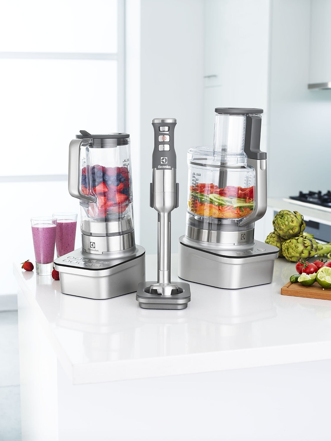 Still life shot of modern kitchen appliances including a blender, immersion blender, and food processor arranged on a clean, white counter.