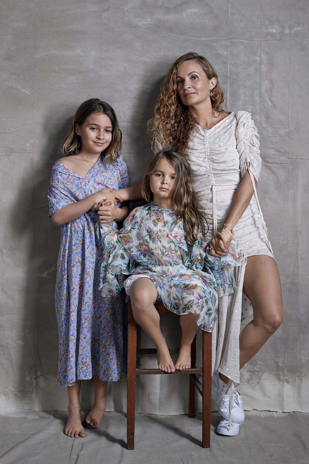 Portrait of a mother with two daughters in floral dresses, one seated on a wooden chair in a brightly lit, neutral studio.