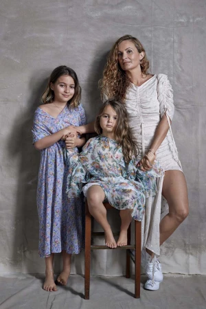 Portrait of a mother with two daughters in floral dresses, one seated on a wooden chair in a brightly lit, neutral studio.