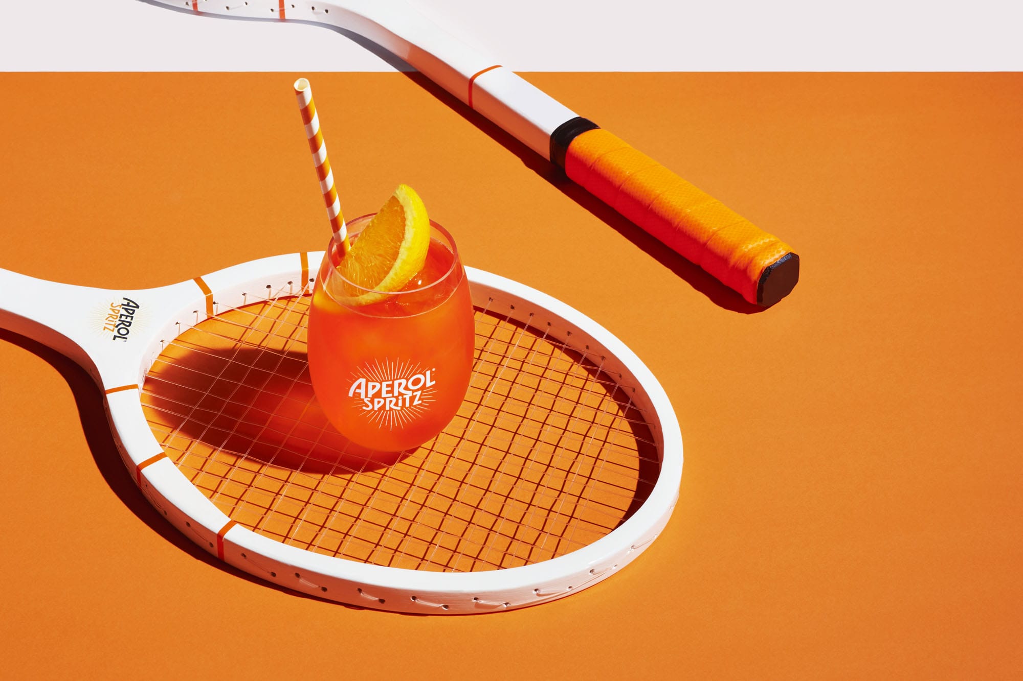 Aperol Spritz cocktail on a tennis racket with an orange slice and straw, set against a bright orange and white background.