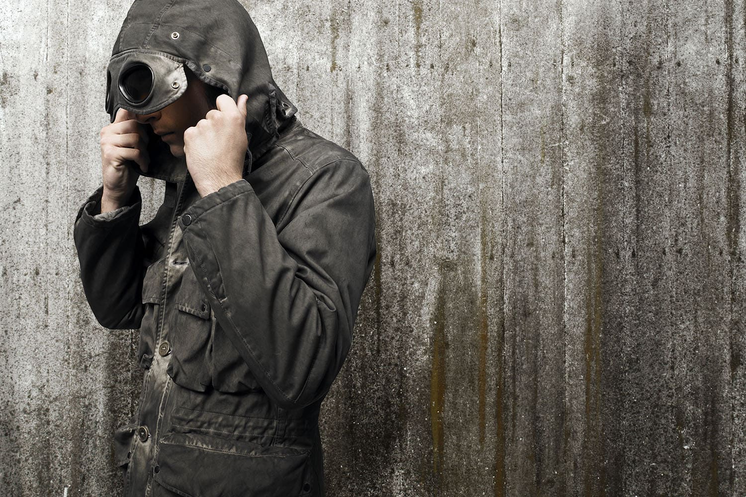 Adult in hooded jacket with goggles stands against textured gray wall. Fashion shot of hooded figure in dark olive green coat with goggle hood.