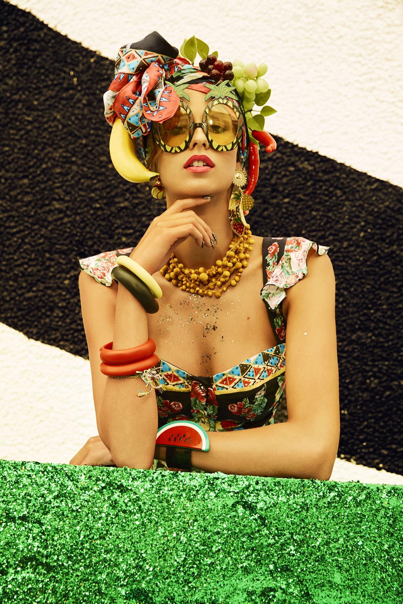 Woman wearing patterned headscarf with fruit and sunglasses, beaded necklace and bracelets, posing against a black and white background.