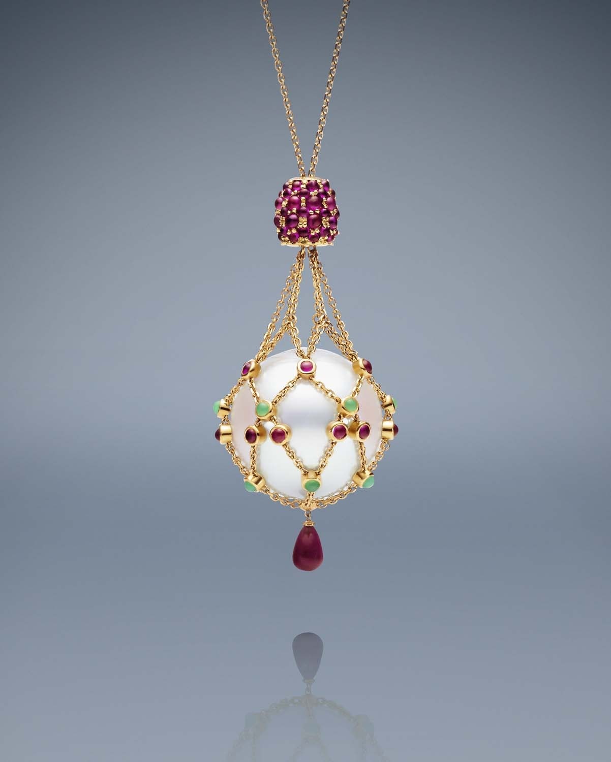 Elegant gold necklace featuring a large white pearl, ruby, and green gemstone details, set against a smooth grey gradient backdrop with reflection.