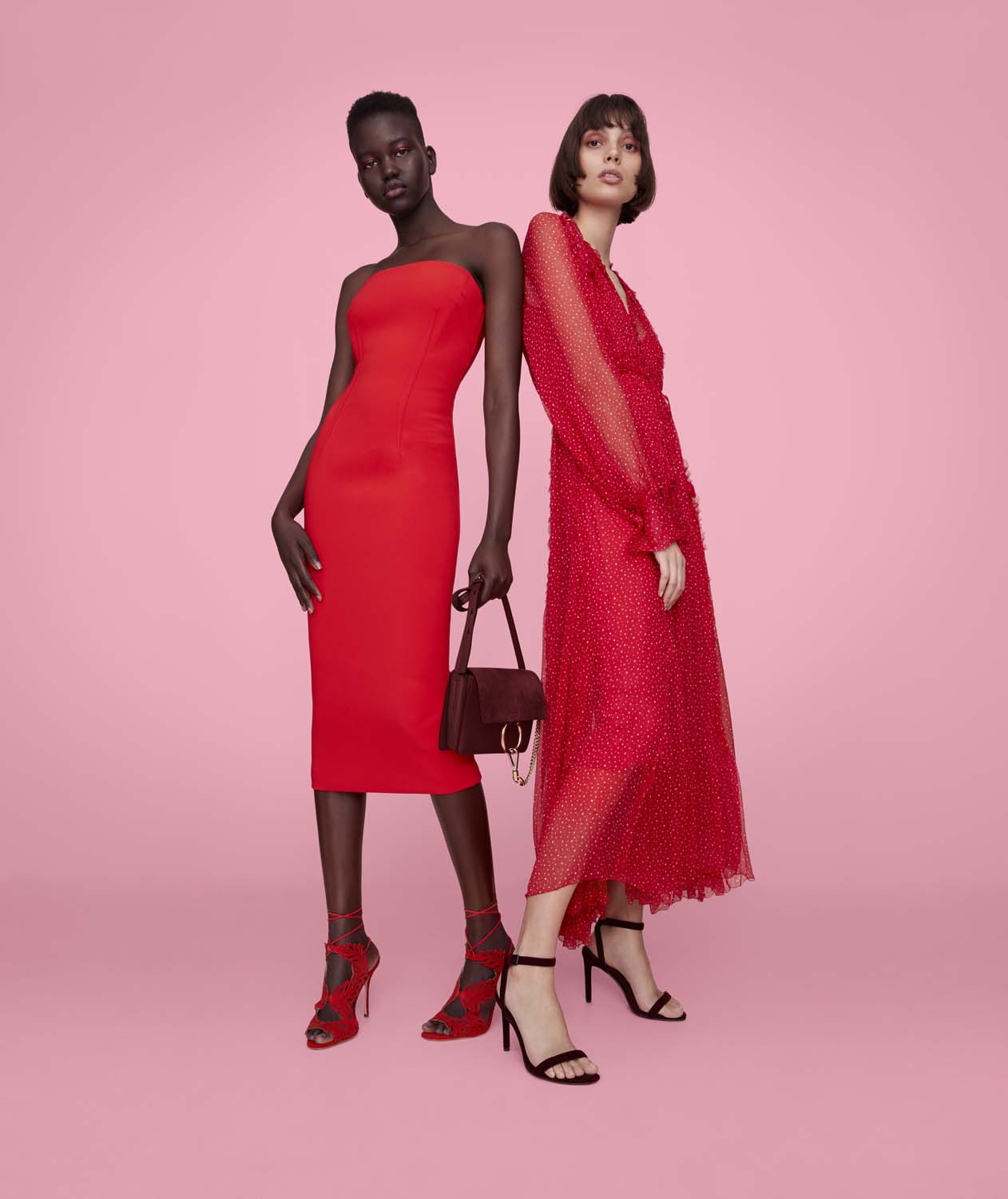 Two adult fashion models in red dresses pose against a pink backdrop, one in a strapless midi dress and the other in a long-sleeved dress.