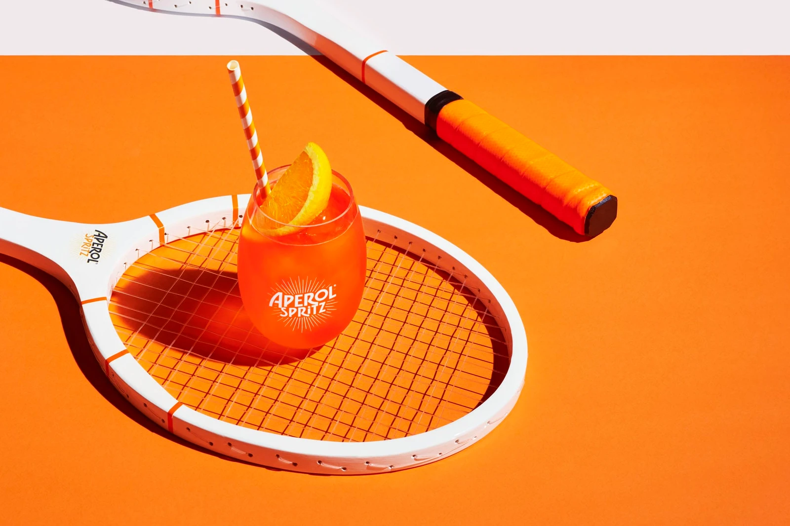 An Aperol Spritz cocktail in a stemless glass with an orange slice and striped straw resting on a tennis racket.