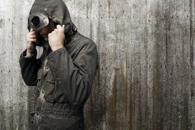 Man wearing a dark hooded jacket with integrated goggles standing against a weathered concrete wall