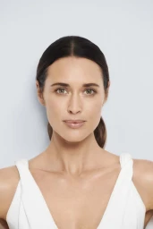 Close up portrait of a woman with dark hair tied back wearing a white top against a light grey background