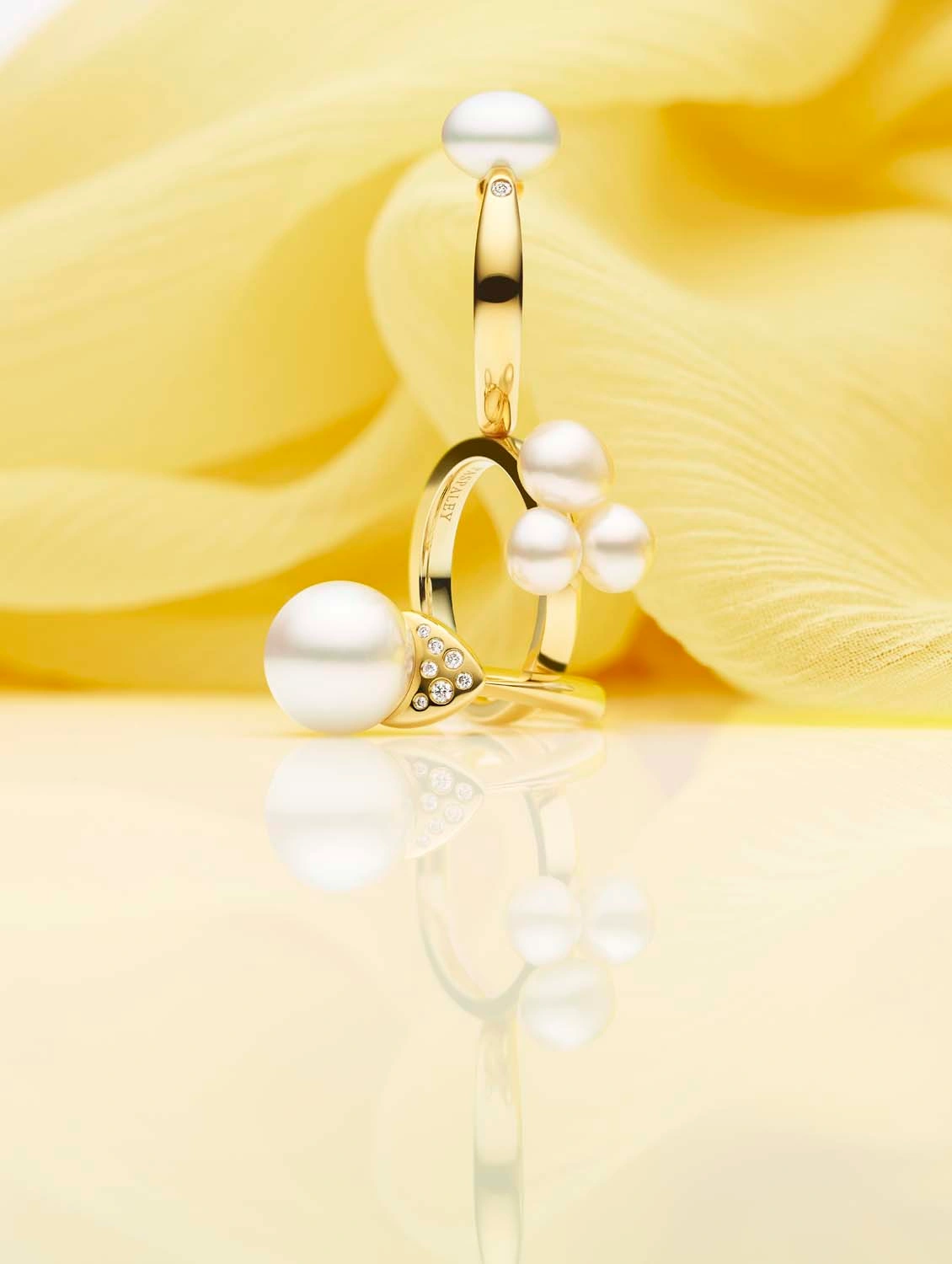 Golden rings adorned with white pearls and small diamonds displayed on a reflective surface against a draped yellow fabric.