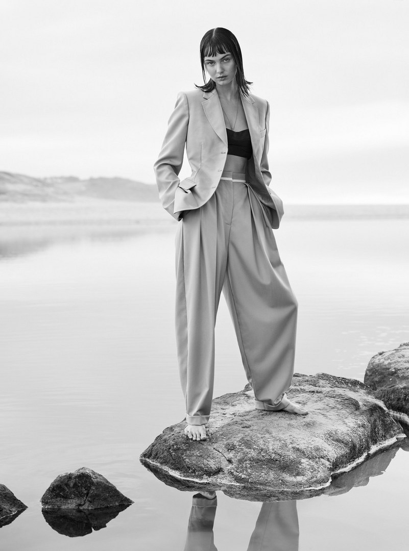 Monochrome portrait of a stylish woman in a suit standing on rocks in shallow water with a hazy horizon in the background.