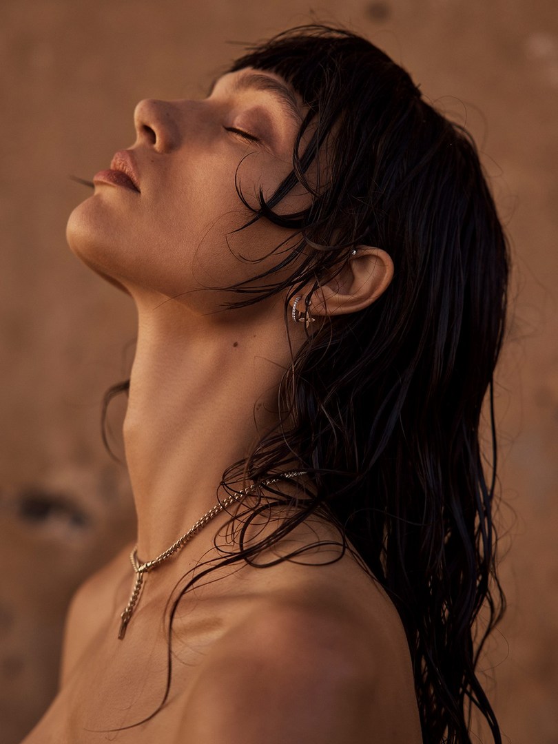 Close-up portrait of a woman with dark hair, eyes closed, wearing a gold necklace, against a textured beige background in soft, warm lighting.