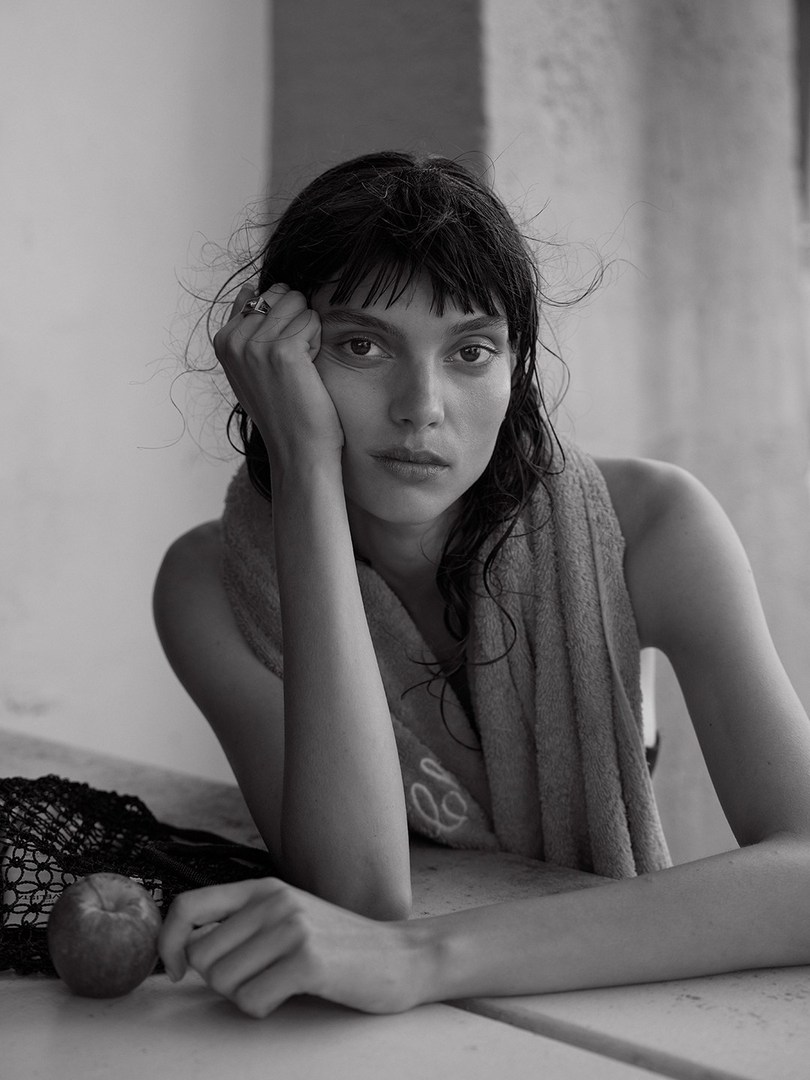 Monochrome portrait of a young woman with wet hair and a towel draped around her neck resting at a table, looking directly at the camera.