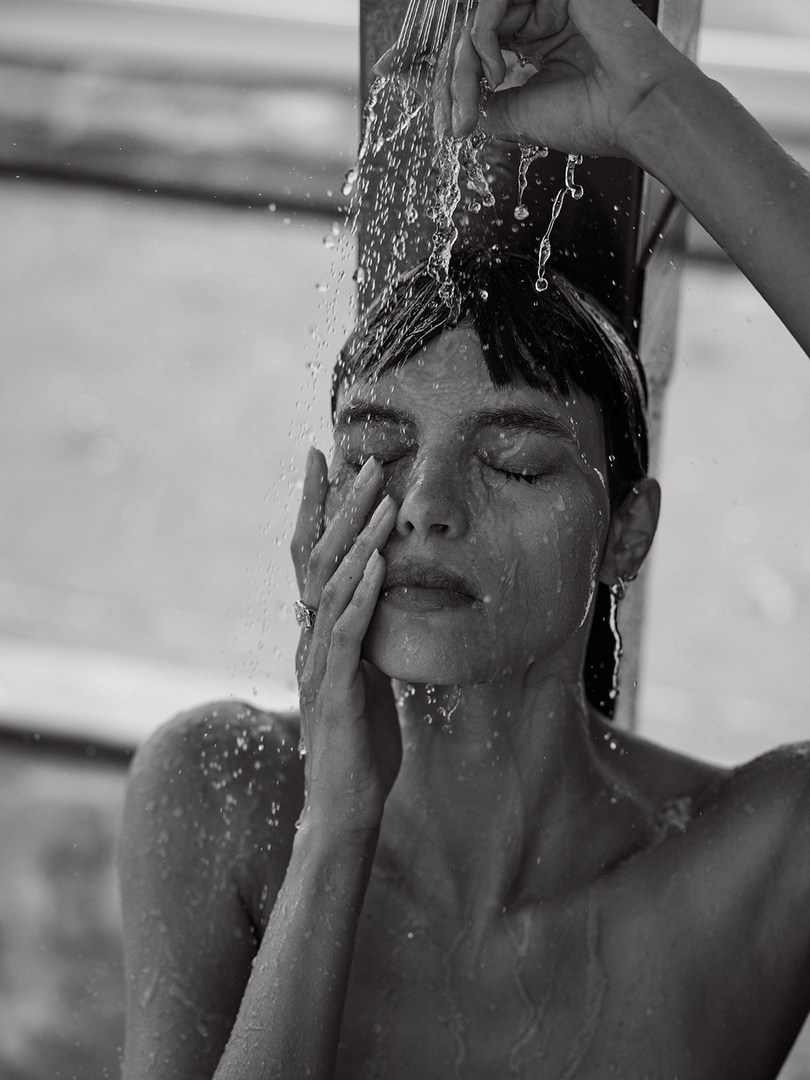 Monochrome portrait of a woman with wet hair and face, eyes closed, standing under a shower, water running over her.