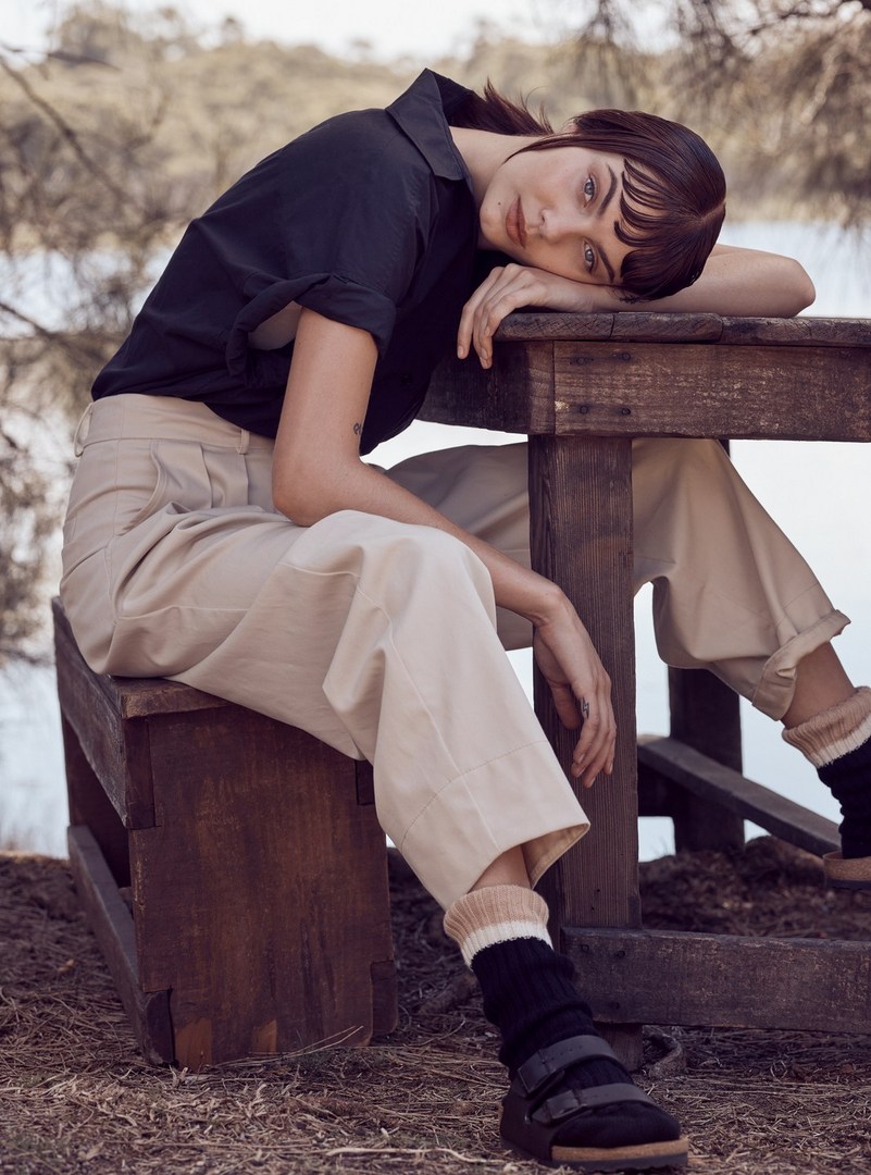 Woman in a black shirt and beige pants poses near a wooden table and bench outside near water and trees in a relaxed outdoor fashion shot.