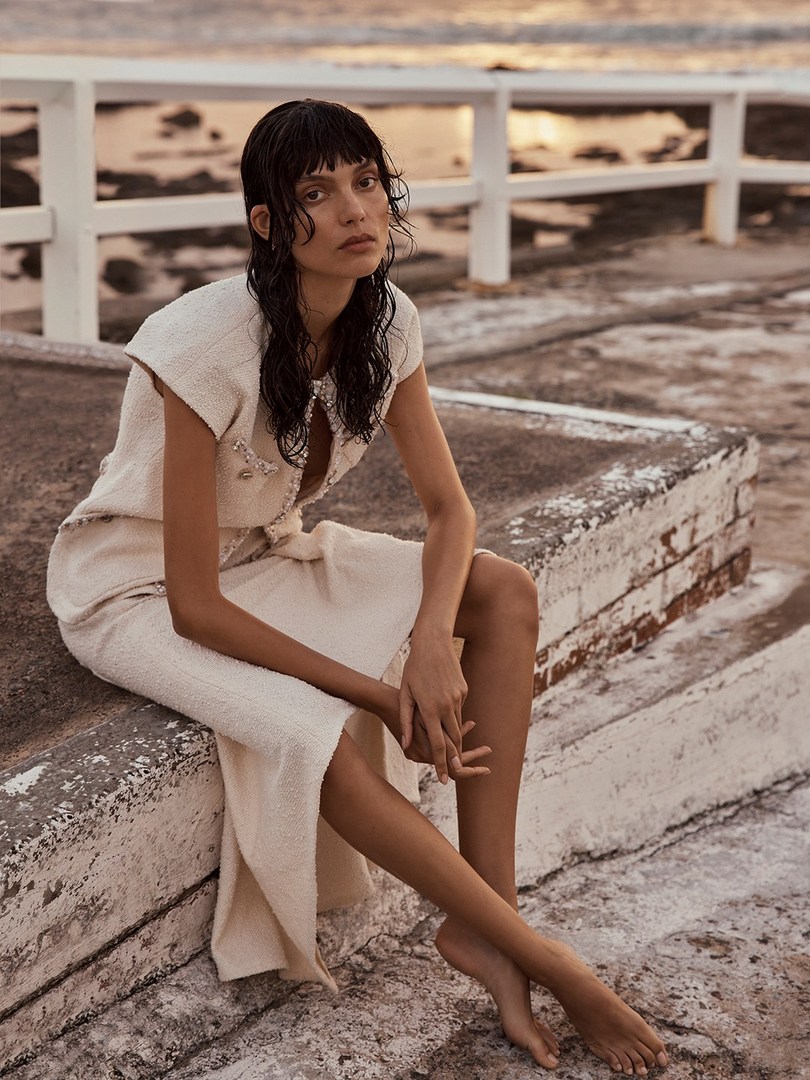 Woman in tweed dress sitting by the ocean at dusk. Cream, beige, and brown tones, with wet hair and bare feet. Fashion photography.