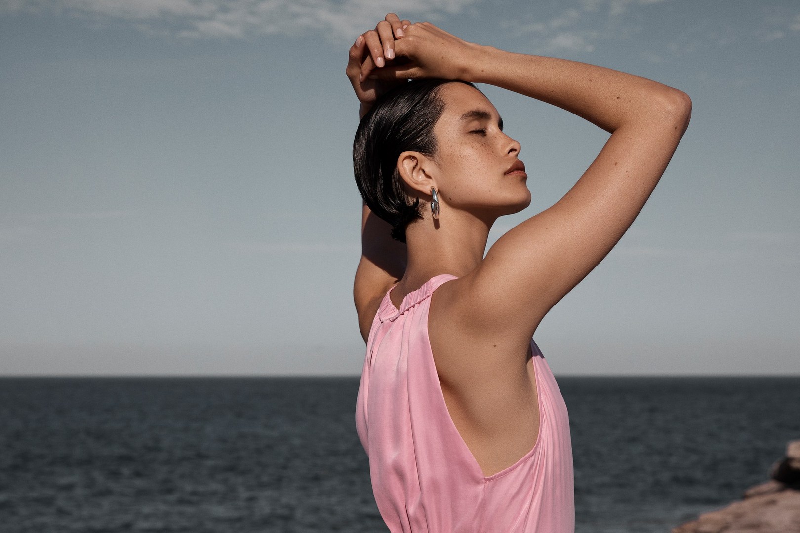 Woman in pink dress with hands behind her head, eyes closed, posing against a coastal background with ocean and sky.