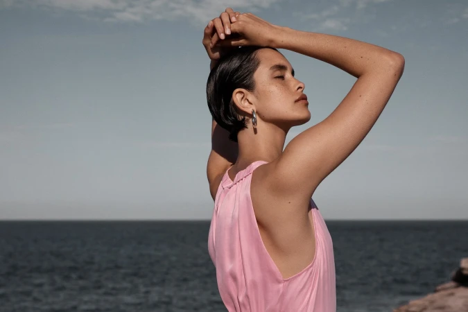 Woman in pink dress with hands behind her head, eyes closed, posing against a coastal background with ocean and sky.