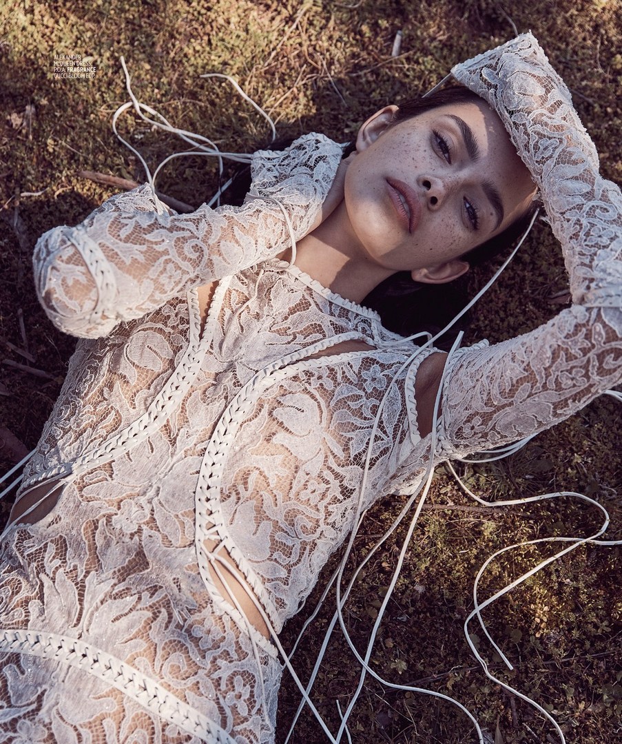 Woman lying on mossy ground wearing white lace dress with hands behind head, looking at camera in eye-level, medium shot.