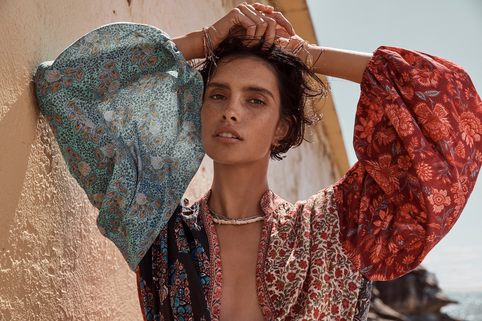 A female fashion model with short hair and a floral-patterned bohemian dress poses against a textured wall in warm, natural light.