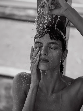 Close-up monochrome portrait of a woman standing beneath a shower with water cascading over her face and dark hair.